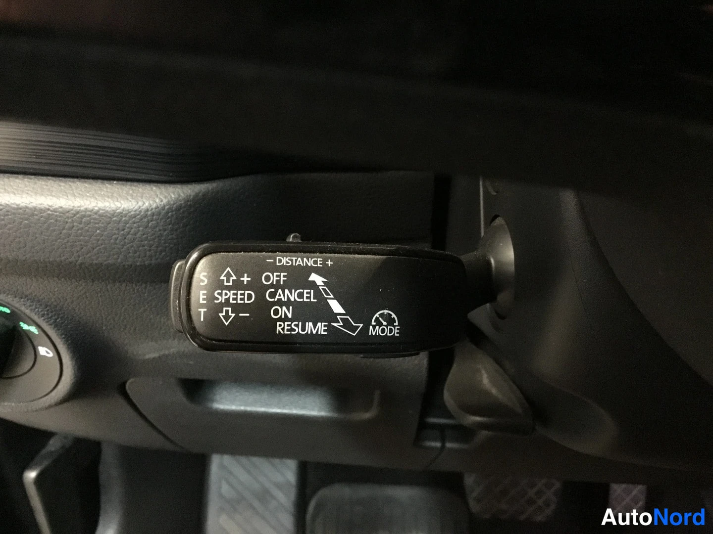 Yksityiskohta: Rear-view camera stalk mounted behind the steering wheel with labels: 'SPEED ON/CANCEL/OFF' and cruise distance settings (+/- DISTANCE) alongside a 'MODE' indic