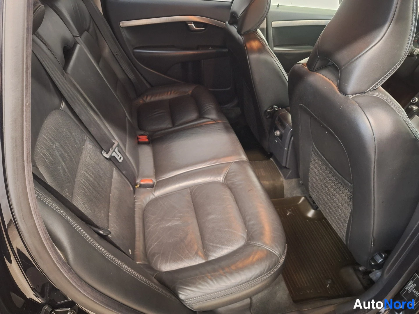 Takaistuimet: View of the rear seating area showing black leather bench seat with three headrests and safety belts. Dark carpeted floor mats and part of the front seat backre
