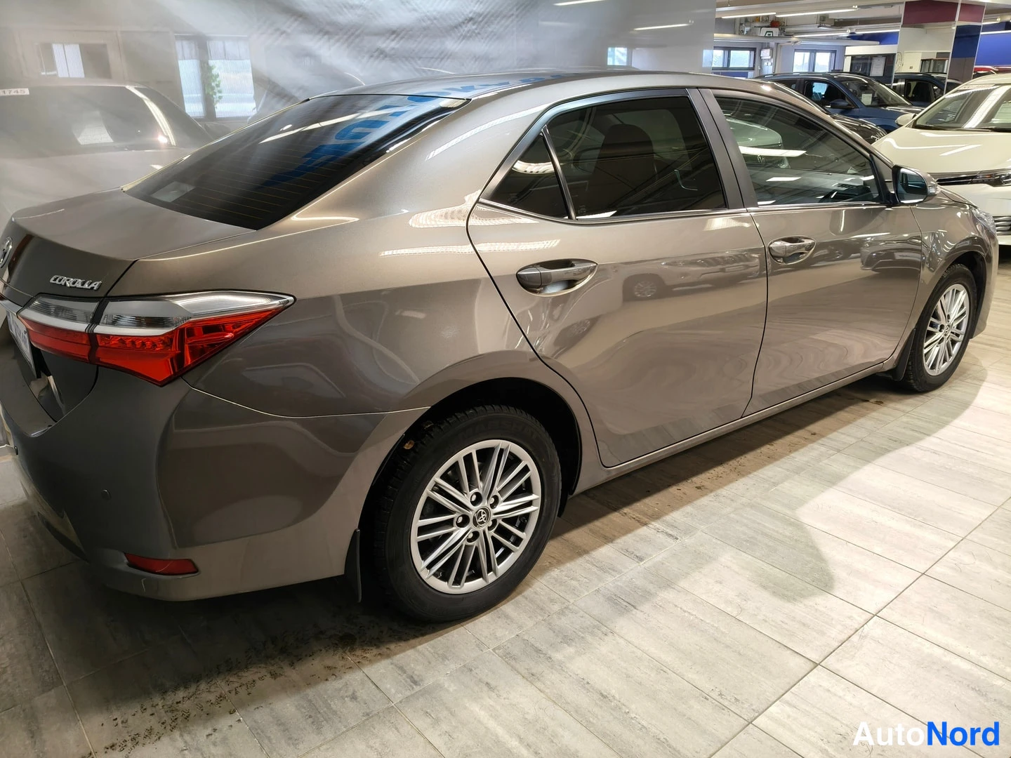 Takaviisto: Takaoikea kolmasosa-kuva hopeanharmaasta Toyota Corolla -sedanista. Takavalo ja 'COROLLA'-nimike näkyvät selvästi. Autossa on kevytmetallivanteet. Koko auto näk