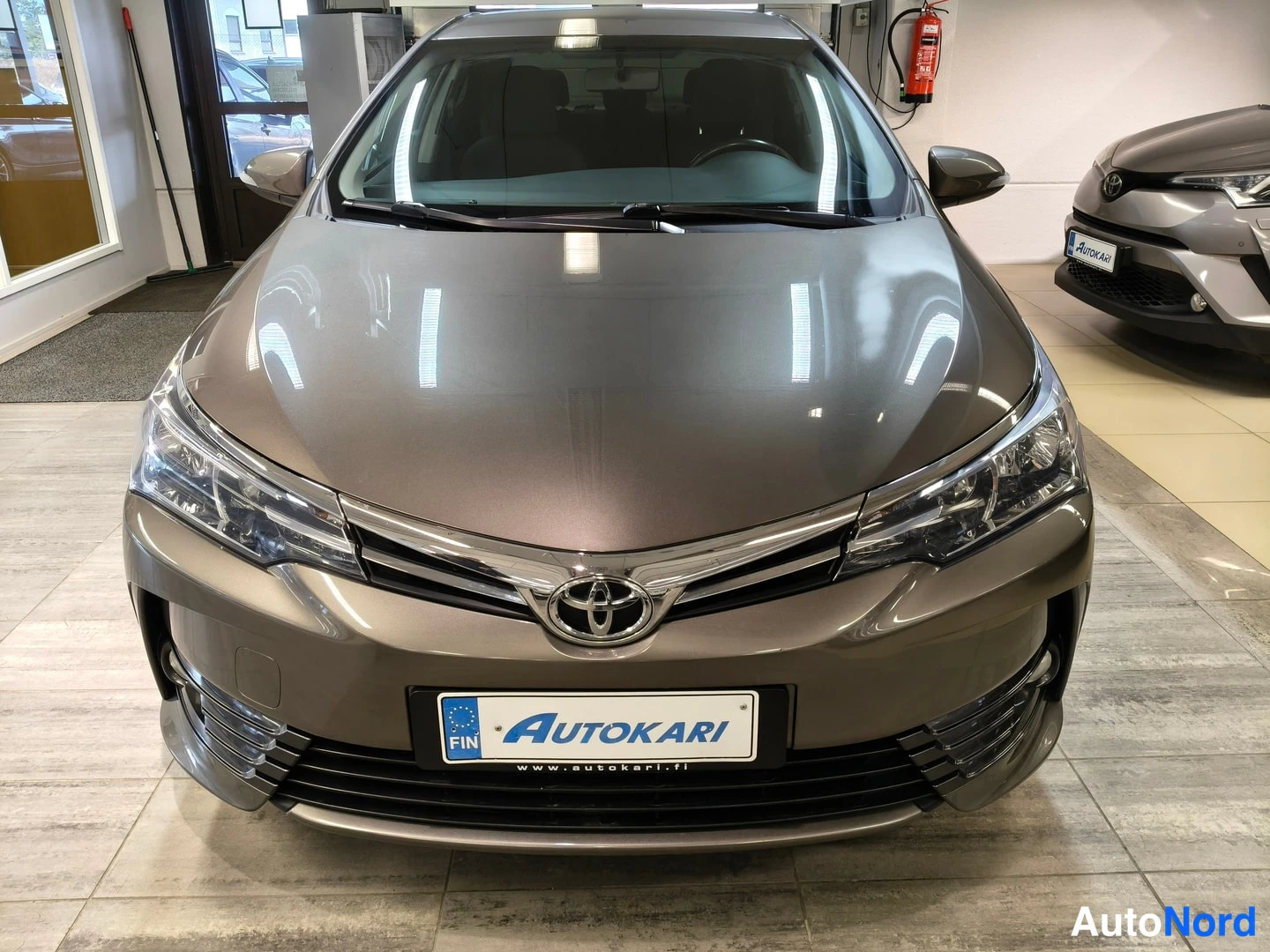 Edestä: Auton keula suoraan edestä: etusäleikössä Toyota-merkki, sumuvalot ja Autokari-tunnuskilpi näkyvät.