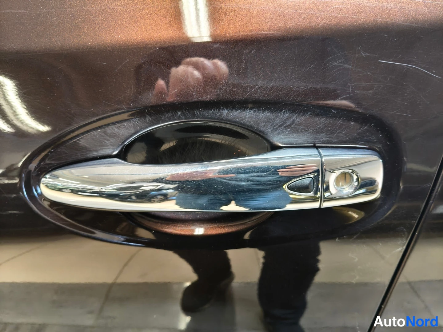 Yksityiskohta: Tarkan kuvan autonosasta, jossa näkyy kromattu ovi-okia pinta, jossa heijastuu kuvaaja ja myyntitila.