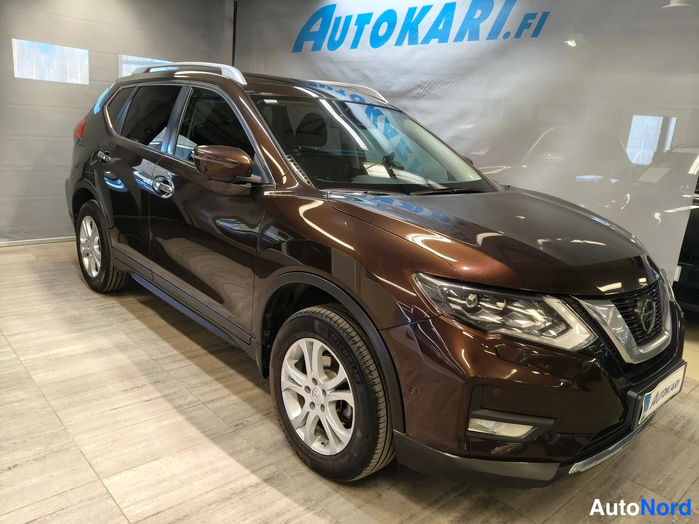 Etuviisto, nokka oikealle: Ruskea Nissan X-Trail kuvattuna kolmianneuvontakuvana oikealta edestä: keula ja vasen kylki näkyvissä, kattoon asennetut hopeiset kaiteet ja hopeat kevyt