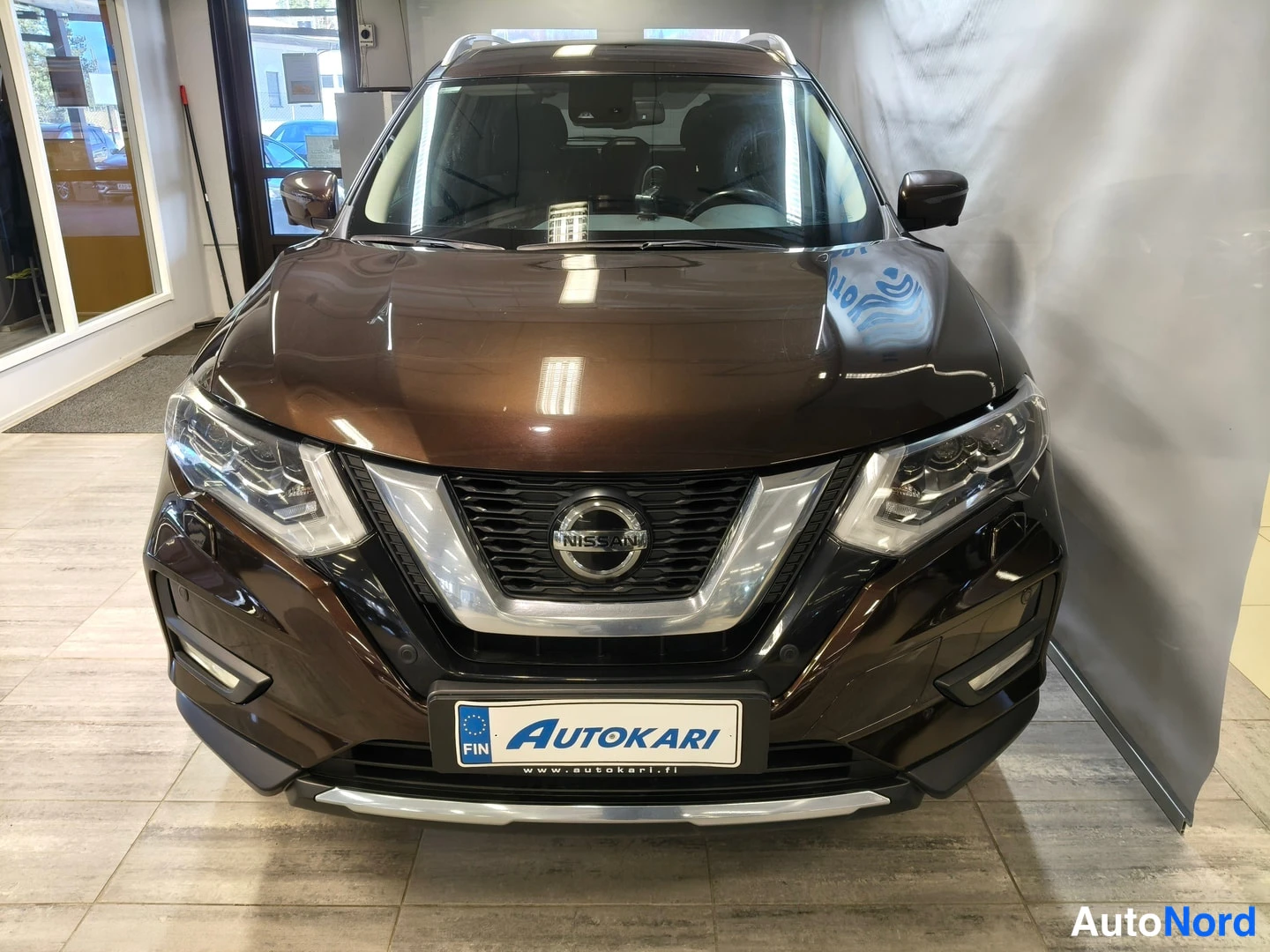 Edestä: Nissan X-Trailin keula suoraan edestä kuvattuna: etusäleikkö ja Nissan-merkki sekä etuvalot näkyvät, konepellillä voimakkaita heijastuksia.