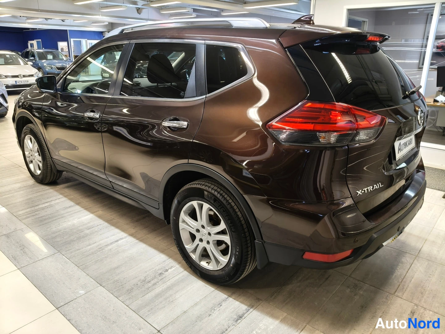 Takaviisto: Ruskea Nissan X-Trail takaa oikealta kuvattuna: takavalot, X-TRAIL-teksti takaluukussa ja vasen kylki näkyvissä, kattoharjat erottuvat.