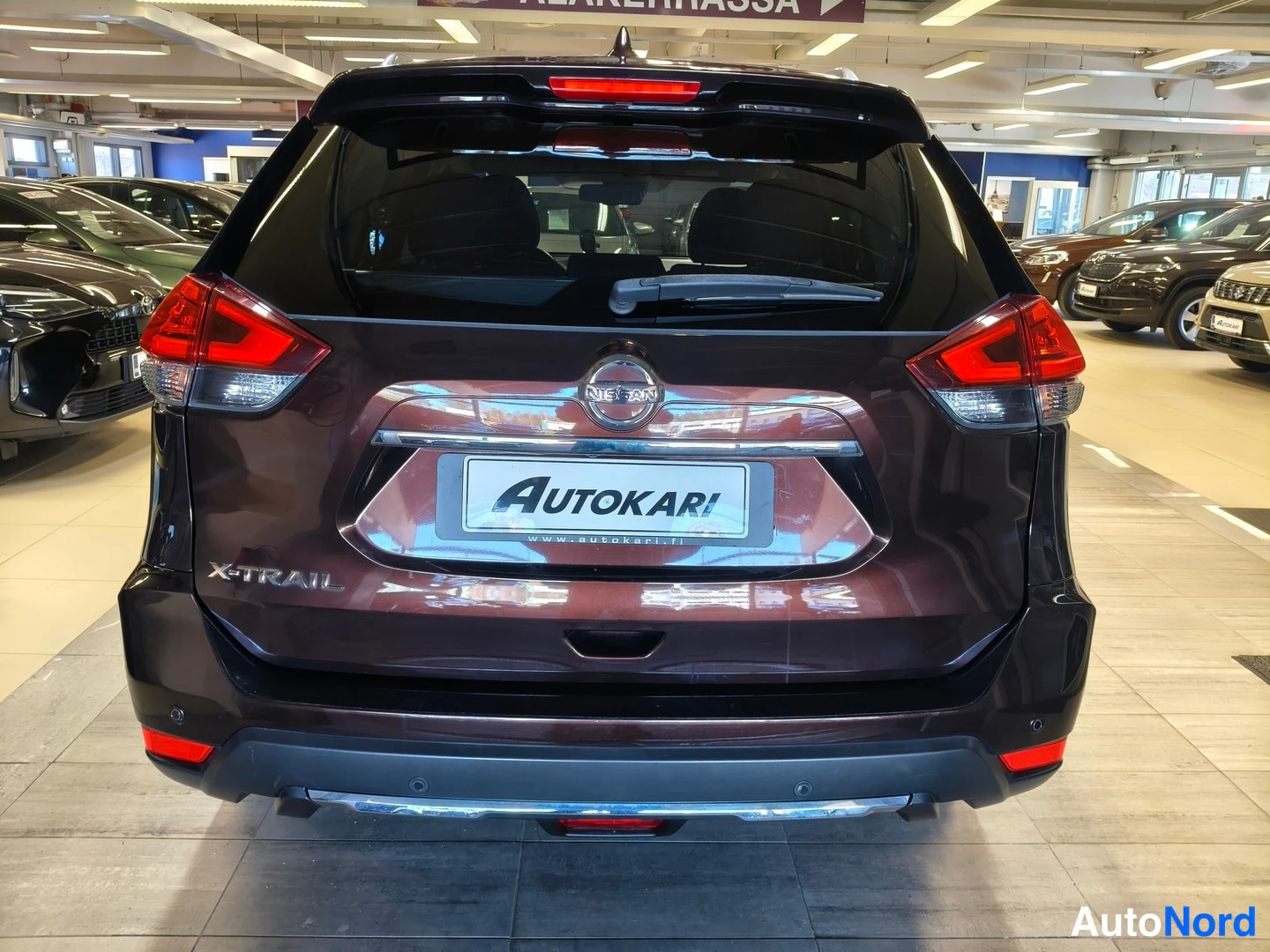 Takaa: Nissan X-Trailin takapää suoraan takaa valaistussa autosalissa: punaiset takavalot, Nissan-merkki ja X-TRAIL-merkki näkyvät, rekisterikilven paikalla AUTOKARI-k