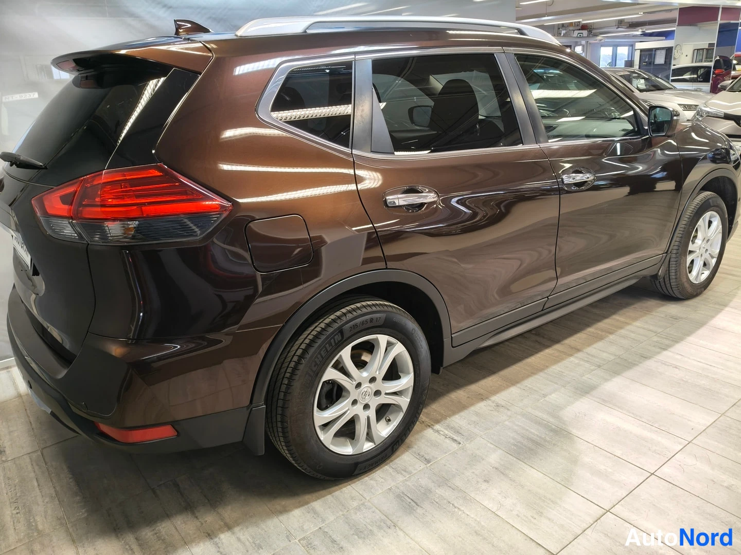 Takaviisto: Nissan X-Trail takaviistosta oikealta: tummanruskea metalliväri, kattokaiteet, hopeiset kevytmetallivanteet ja takavalot näkyvät autosalin lattialla.