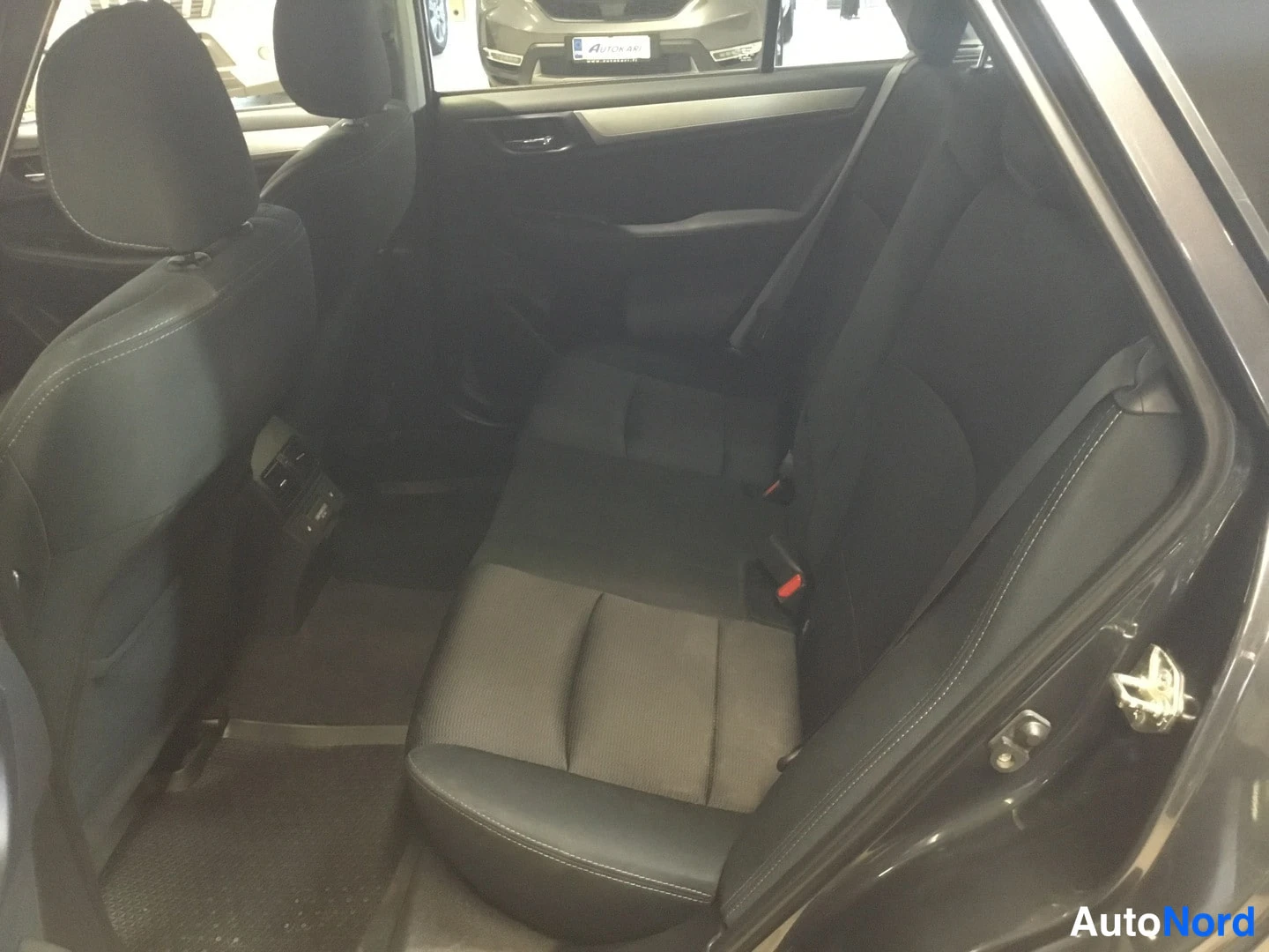 Takaistuimet: Rear passenger compartment showing black fabric rear seats, a floor mat, and the back of the front seat. The driver-side door is open.
