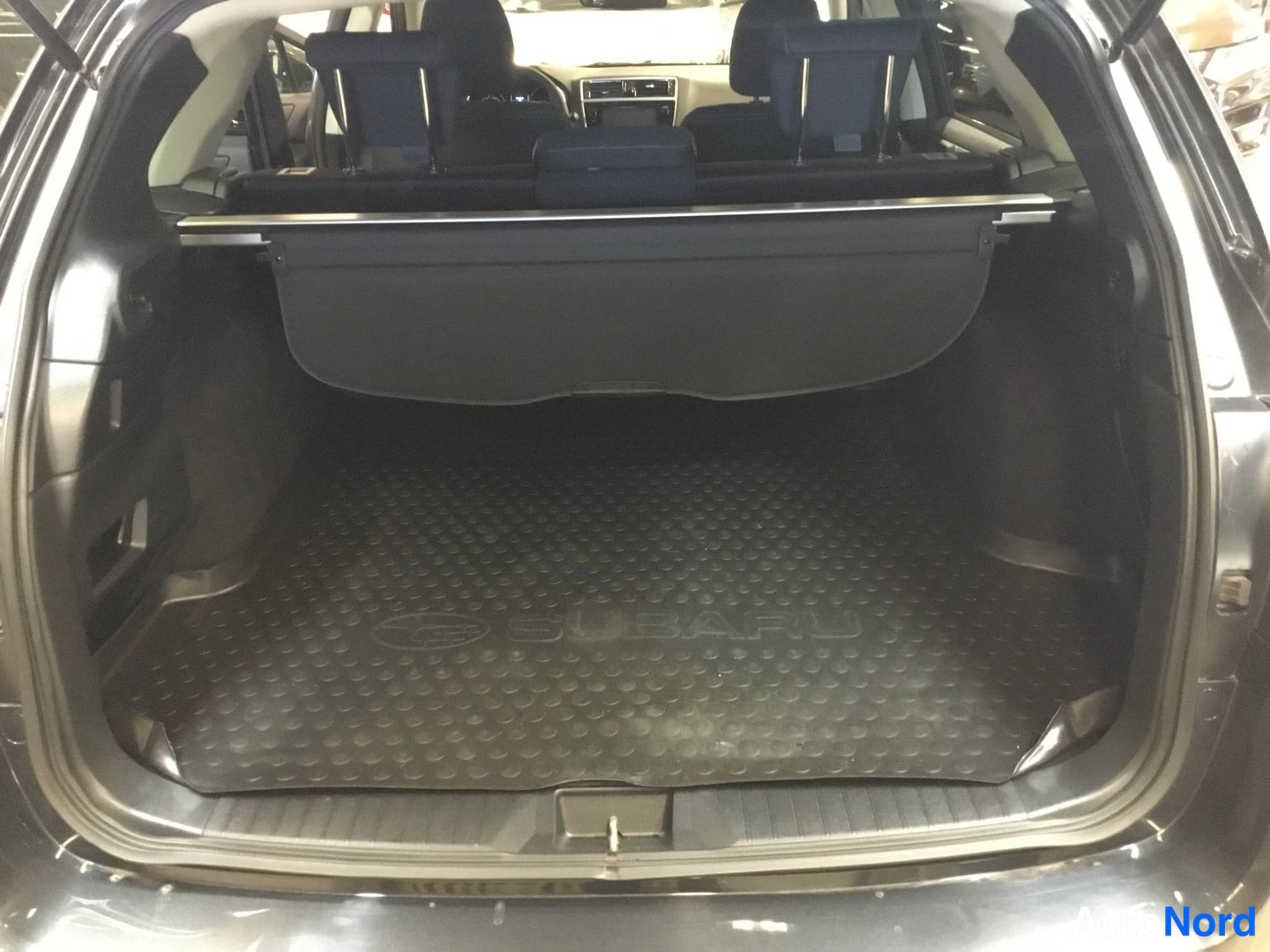 Tavaratila: Open trunk with black plastic floor mat featuring embossed Subaru logo and oval emblem. Retractable cargo cover positioned behind rear seats.