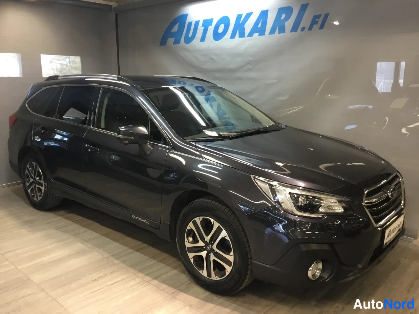 Etuviisto, nokka oikealle: Subaru Outback -farmari tummanharmaana etuviistossa vasemmalta, kotelo ja luvut näkyvät Autokari-showroomissa.