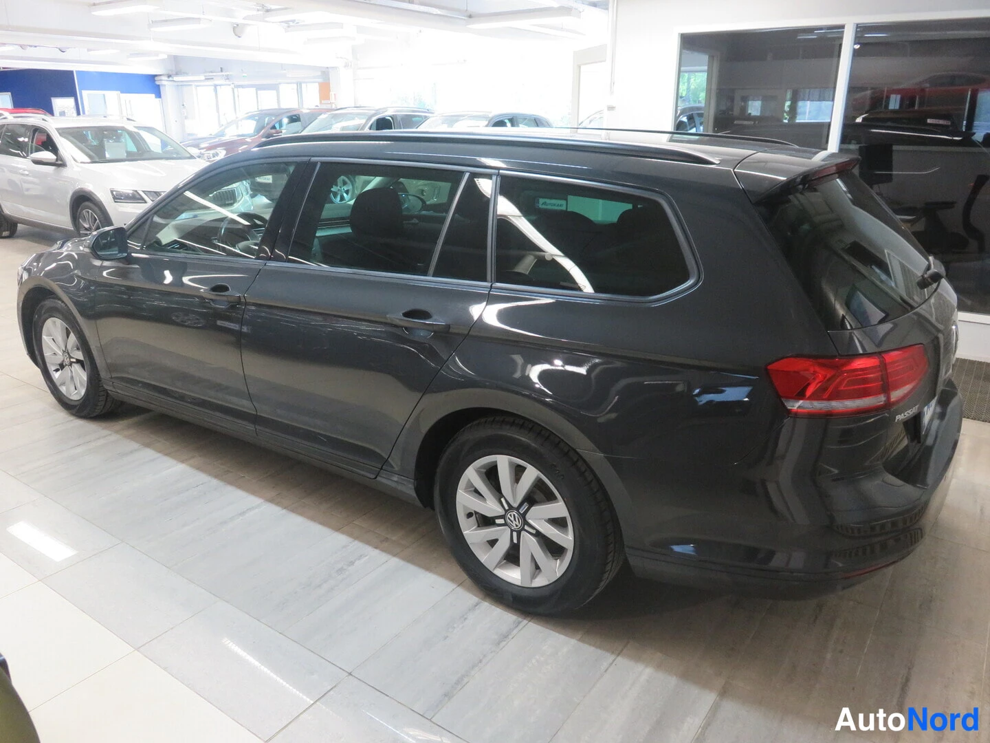 Takaviisto: Rear three-quarter view of a dark grey station wagon inside a showroom. Roof rails and alloy wheels are visible.