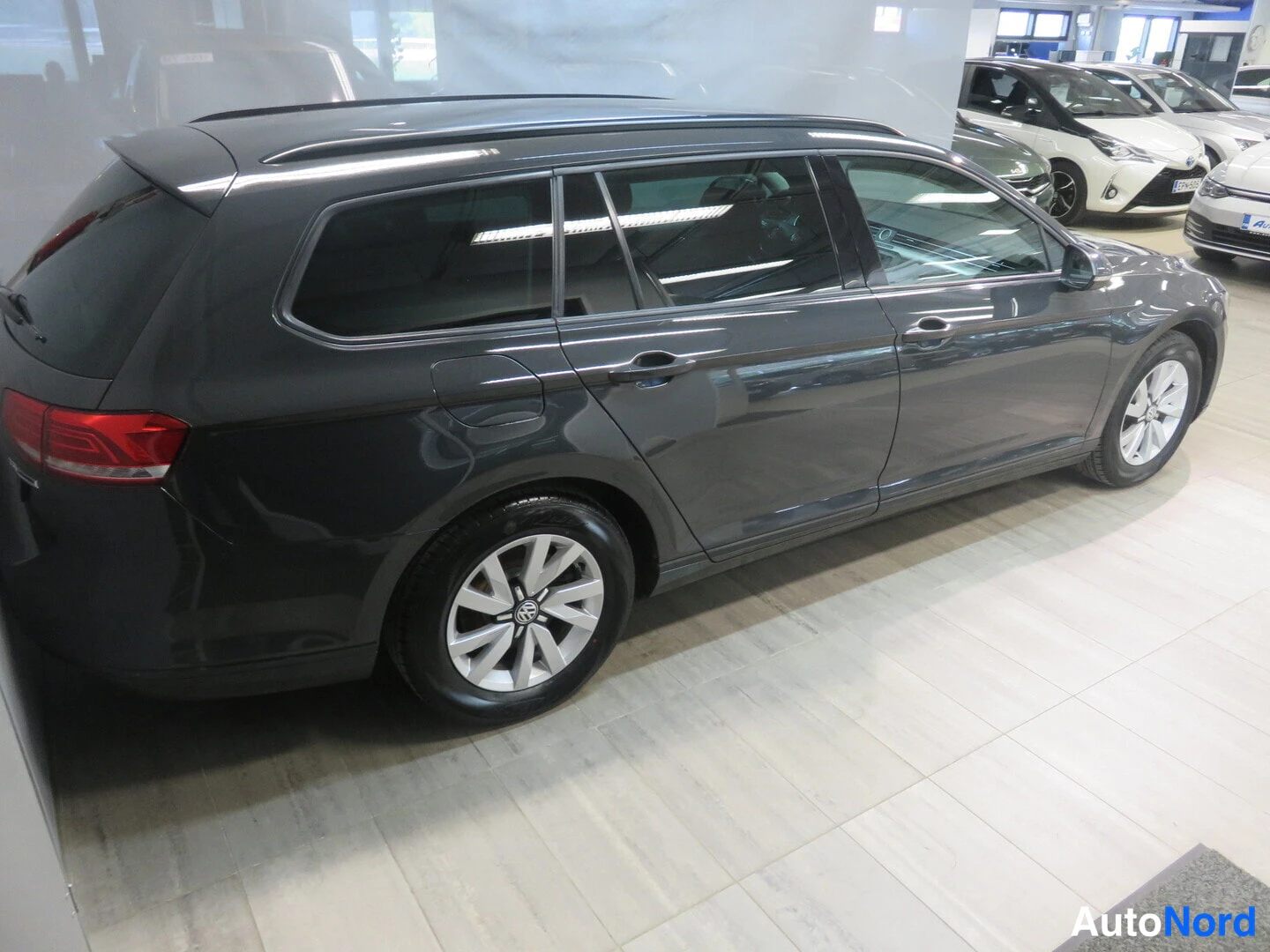 Sivulta: Side view of the dark grey station wagon parked in a showroom. Alloy wheels and roof rails are clearly visible.