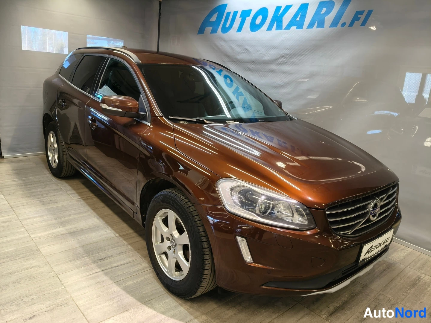 Etuviisto, nokka oikealle: Brown Volvo SUV from front three-quarter view inside showroom with 'AUTOKARI.FI' on back wall.