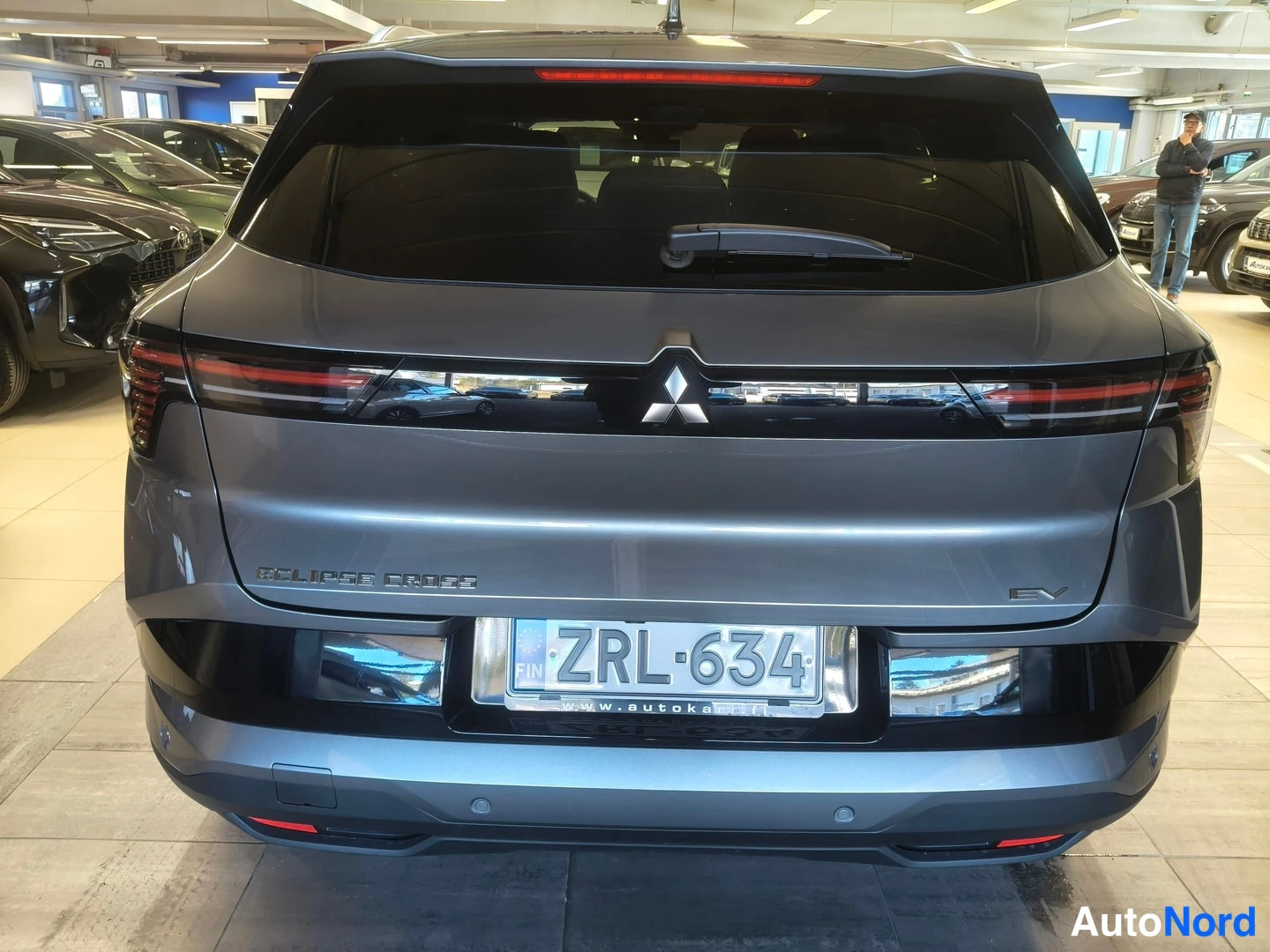Takaa: Auton takaosa: Mitsubishi Eclipse Cross -merkintä ja EV-merkintä takaluukussa, rekisterikilpi, viistoperä harmaana sisätiloissa.