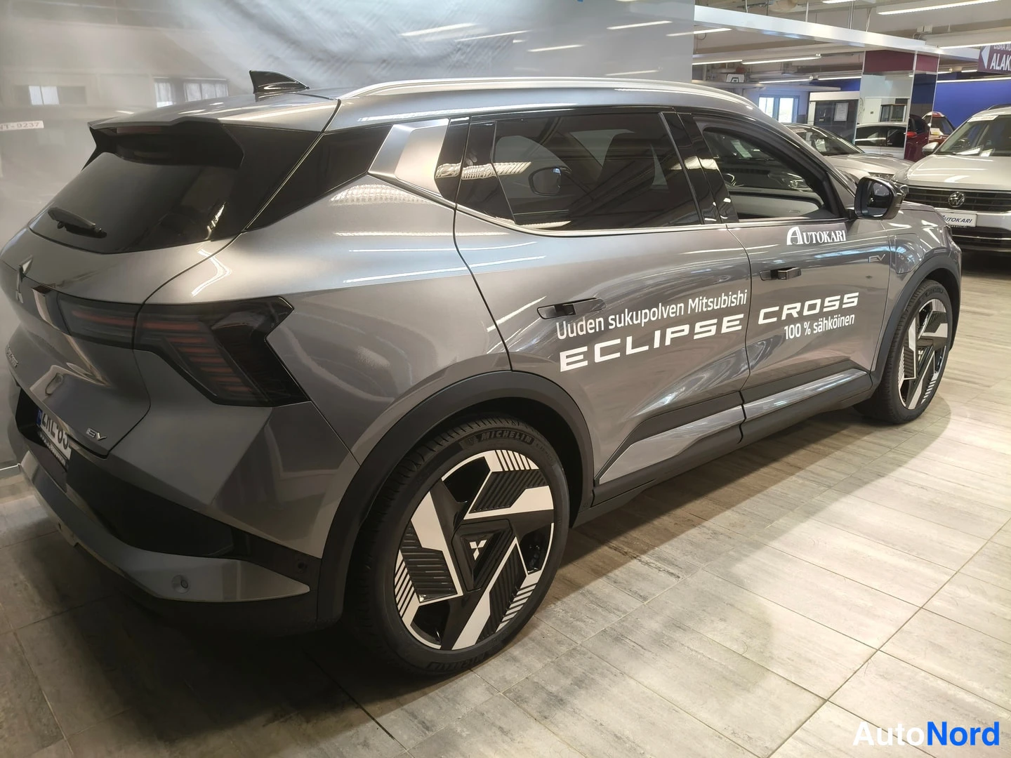 Takaviisto: Auton takaosa ja oikea kylki kolmannelta sivulta: harmaa Mitsubishi Eclipse Cross, kyljessä teksti 'Uuden sukupolven Mitsubishi ECLIPSE CROSS 100% sähköinen', A