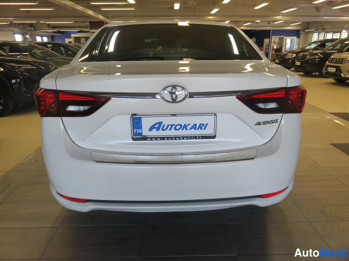 Takaa: Valkoisen sedanin takapää; peräkontissa kyltti Autokari ja merkintä AVENSIS.