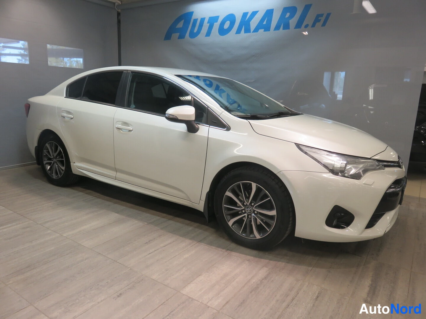 Etuviisto, nokka oikealle: Valkoinen Toyota Avensis -sedan etukulmasta oikealta sisätiloissa.