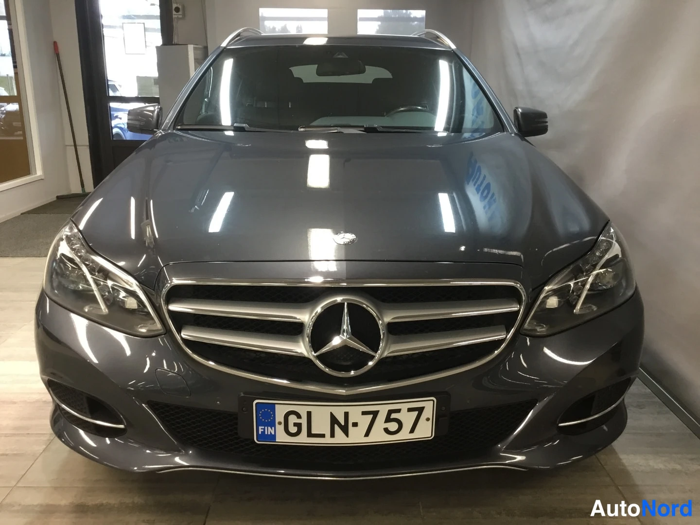 Edestä: Mercedes-Benz farmarin tummanharmaa etupäää näkyvissä etuoven levikkeineen; kylki- ja merkistökiinnitys selkeästi näkyvissä.
