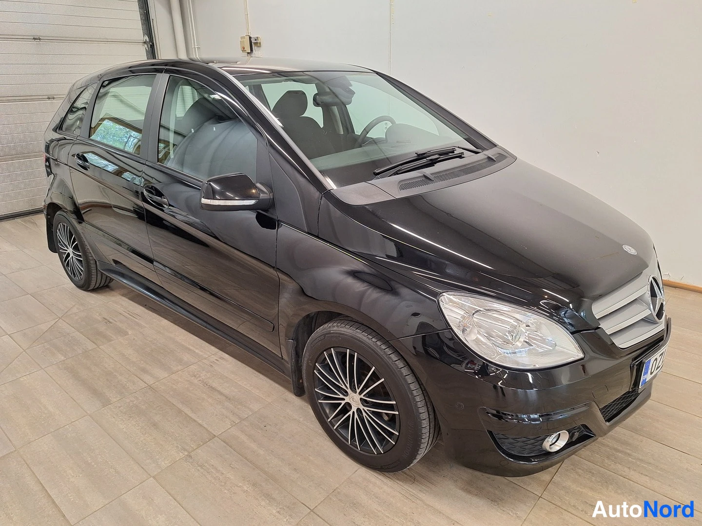 Etuviisto, nokka oikealle: Musta Mercedes-Benz B 180 etu-neljännesvasen kuvakulma sisätiloissa, keulan ja molempien sivujen näkyvissä.