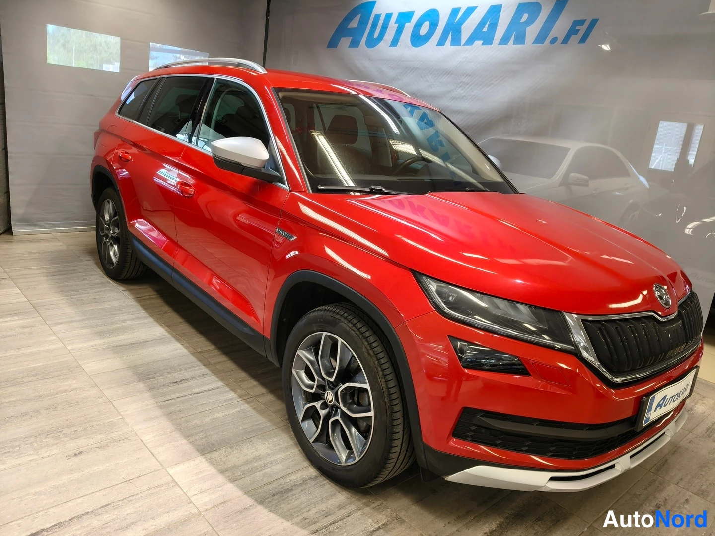Etuviisto, nokka oikealle: Punainen Skoda Kodiaq esitellään showroomissa etukulmasta oikealta; etusäleikkö, ajovalot ja alumiinivanteet näkyvät.