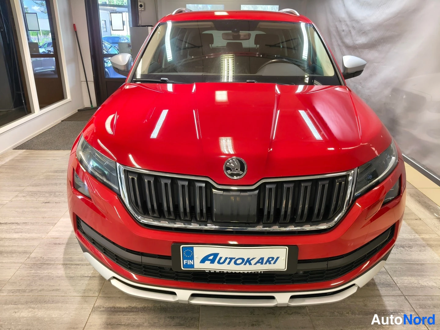 Edestä: Etukatselu punaisesta Skoda Kodiaqista showroomissa; merkkiäinen etusäleikkö, ajovalot ja Autokari-tarra etupuskurissa.