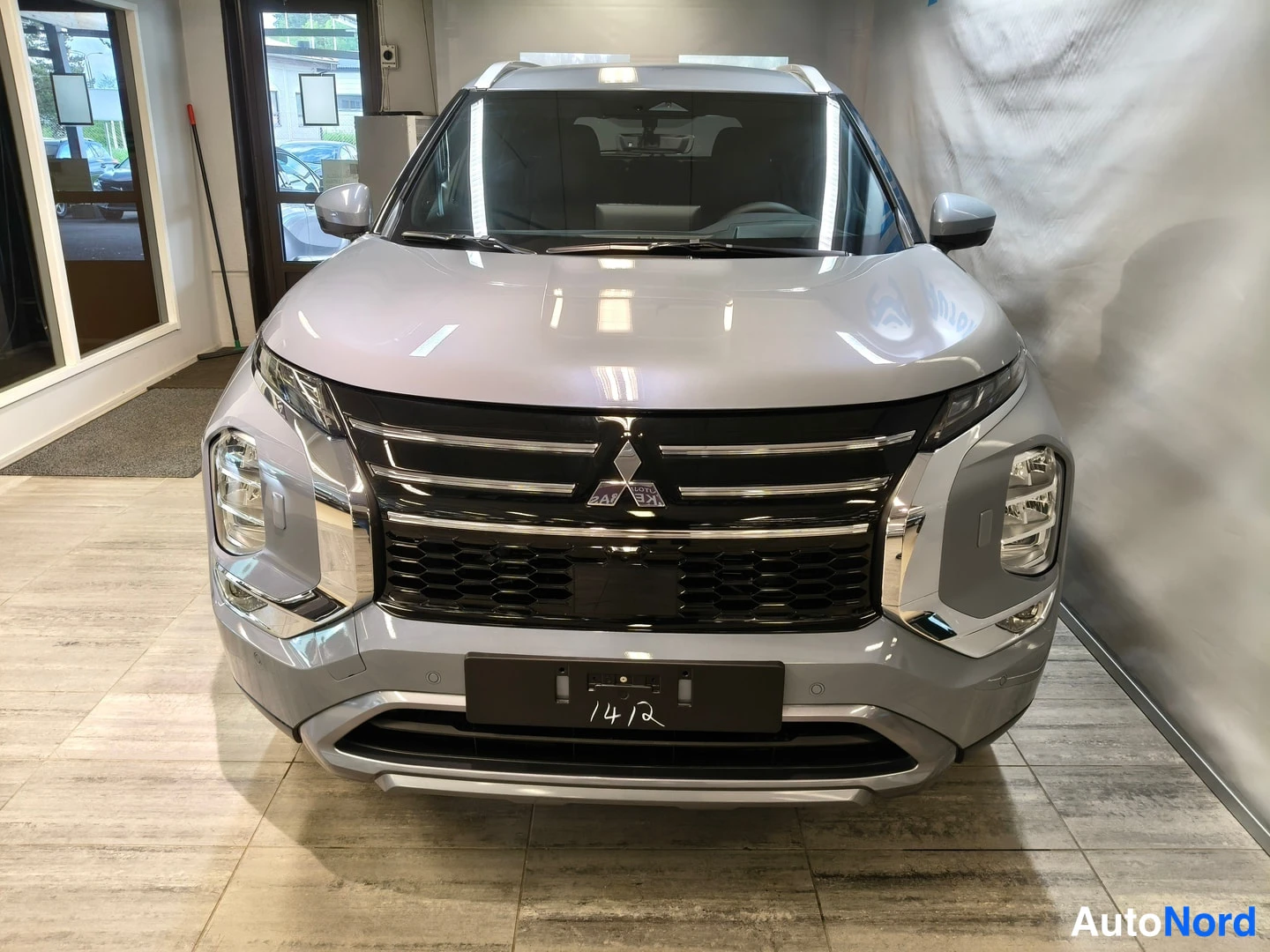 Edestä: Suora etukuva hopeisesta autoista sisätiloissa. Auton pinnalla on heijastuksia. Eturitilässä on selkeästi Mitsubishi-malli. Pistorasia-merkki ei ole kuvassa näk