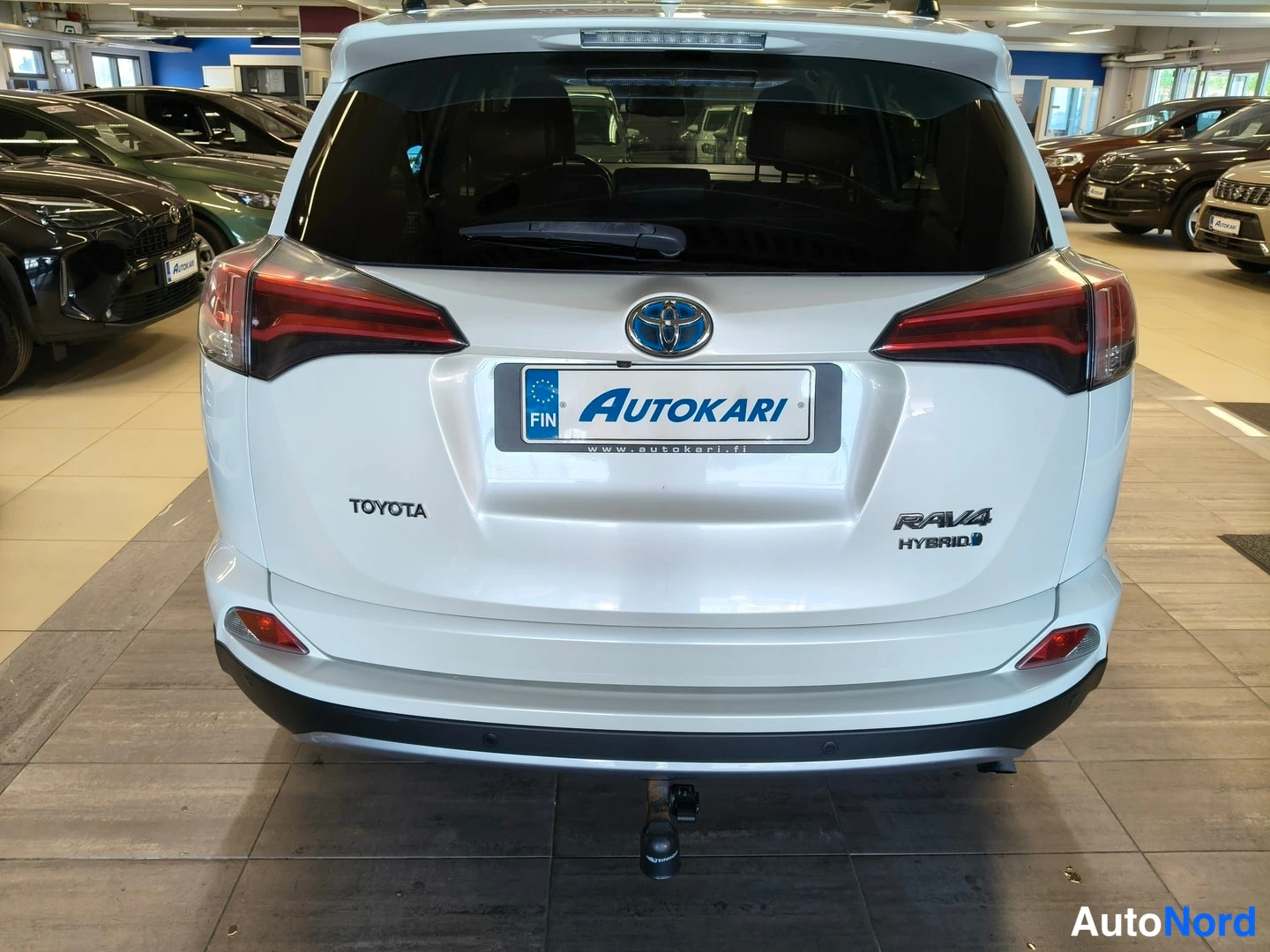 Takaa: Auton takanäkymä: valkoinen Toyota RAV4 Hybrid -merkinnöin, Autokari-kilpi ja kytketty vetokoukku autosalon lattialla.