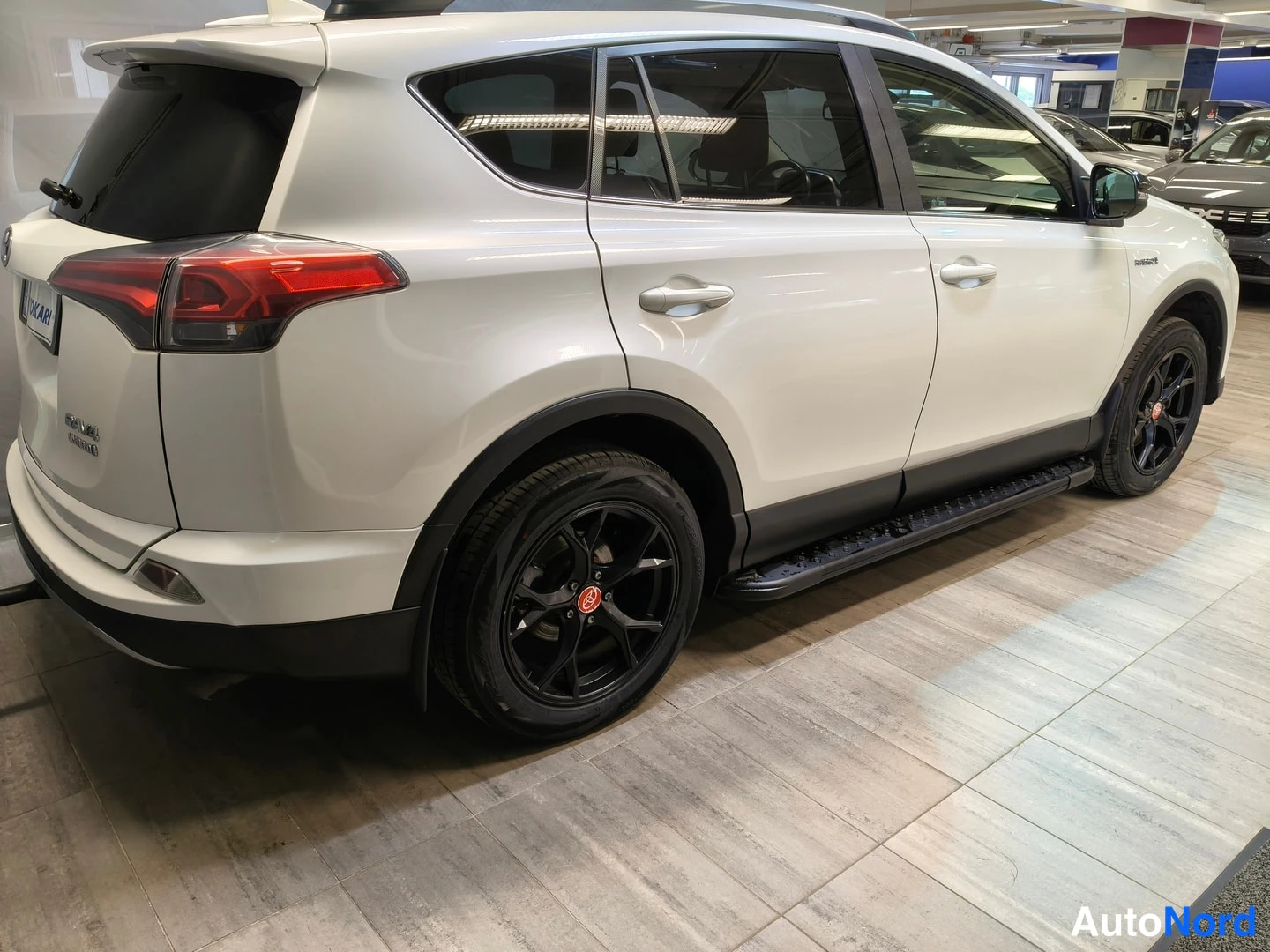 Takaviisto: Taka sivukulmanäkymä valkoisesta Toyota RAV4 Hybristä: mustat kevytmetallivanteet ja sivukaiteet, autotallin tiilikiveys lattialla.