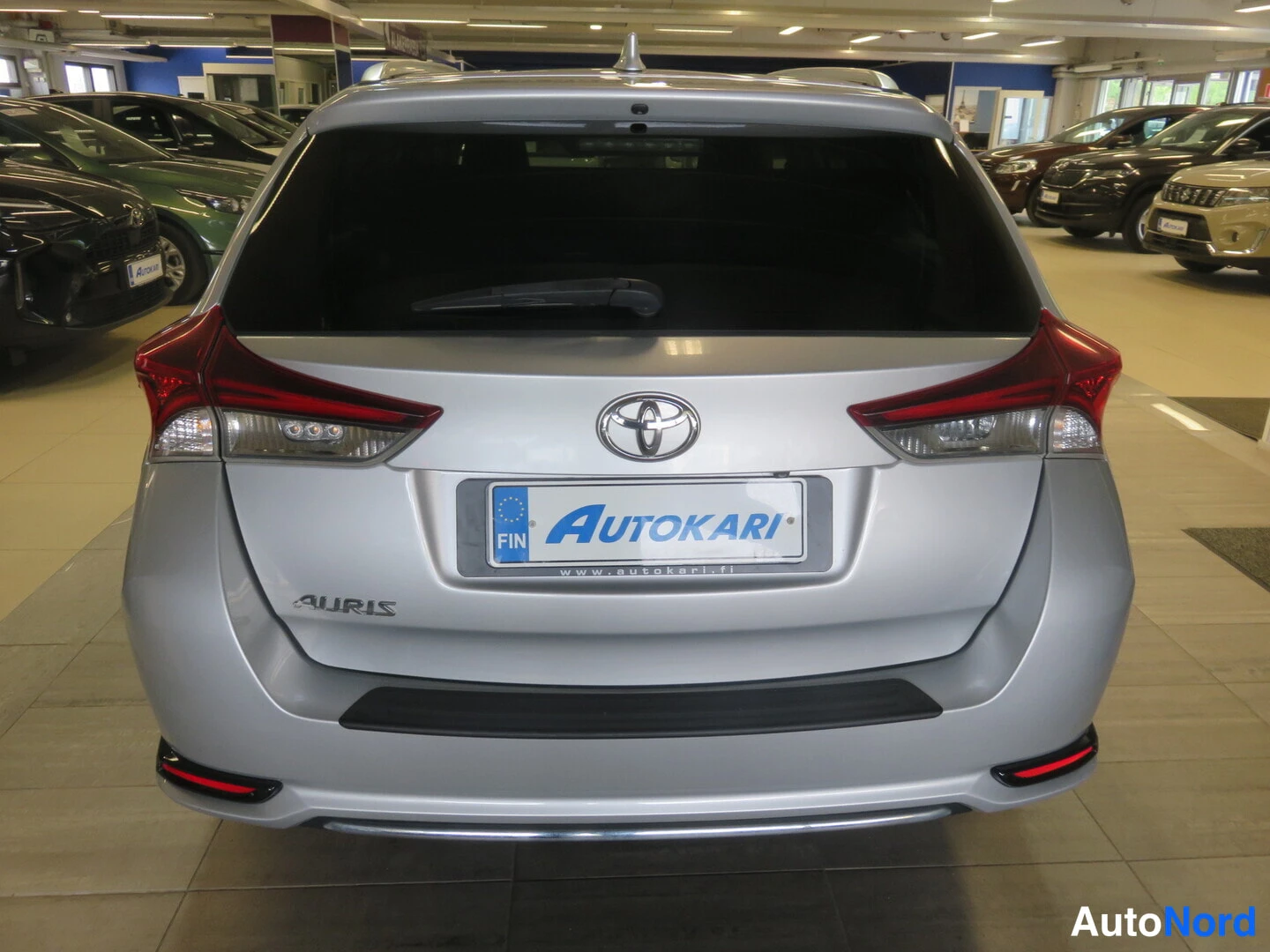 Takaa: Hopeanharmaan Toyota Auriksen takapää: punaiset takavalot, Toyota-merkki ja Auris-teksti, Autokari-kyltti.