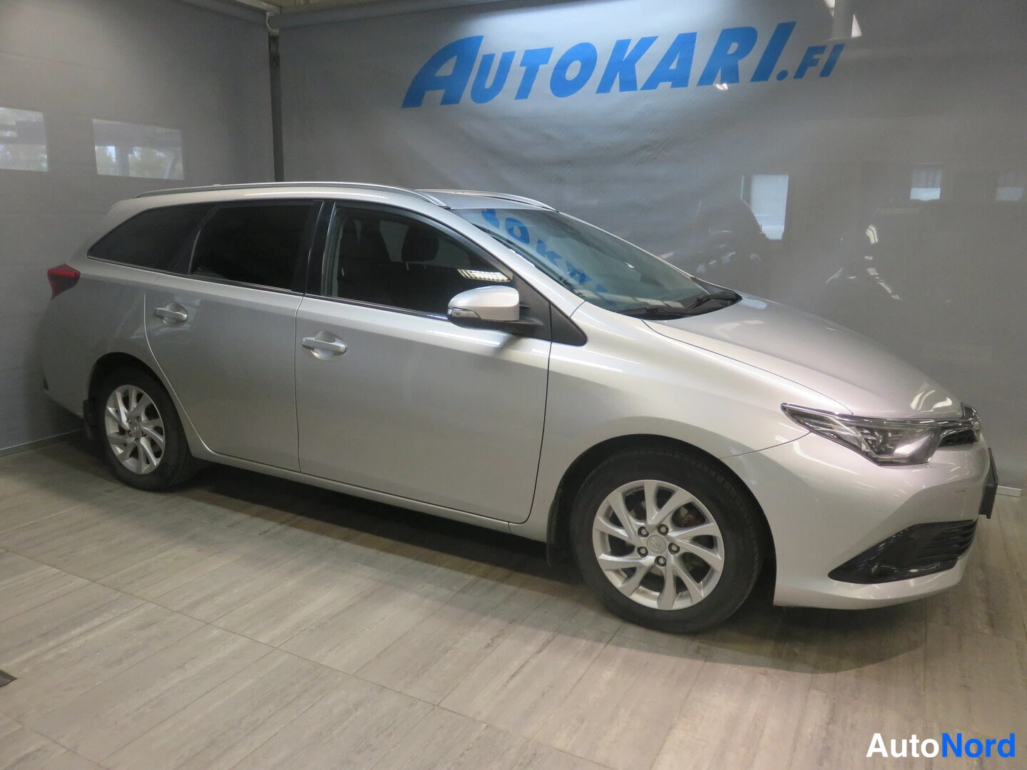 Etuviisto, nokka oikealle: Hopeanvärinen Toyota Auris Touring Sports -farmariauto kuvattuna kolmasosakulmasta keulaa oikealle taakse seinätessä showroomissa