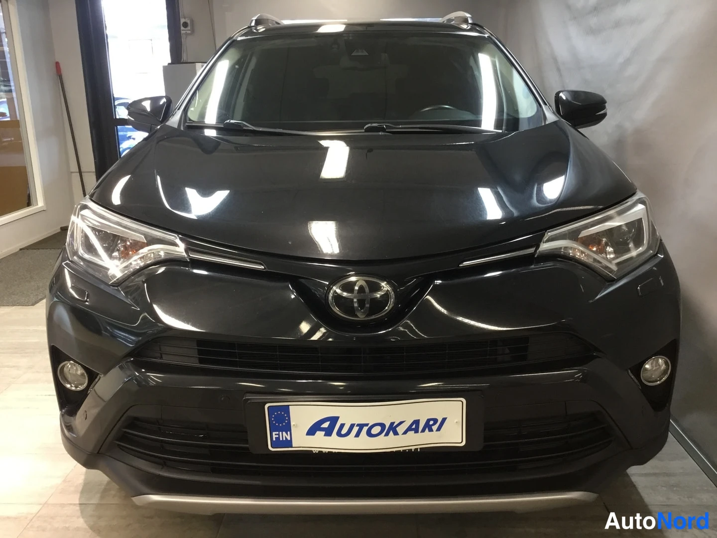Edestä: Mustan Toyota RAV4:n etunäkymä showroomissa, Toyota-merkki ja Autokari-kilpi etupuskurissa.