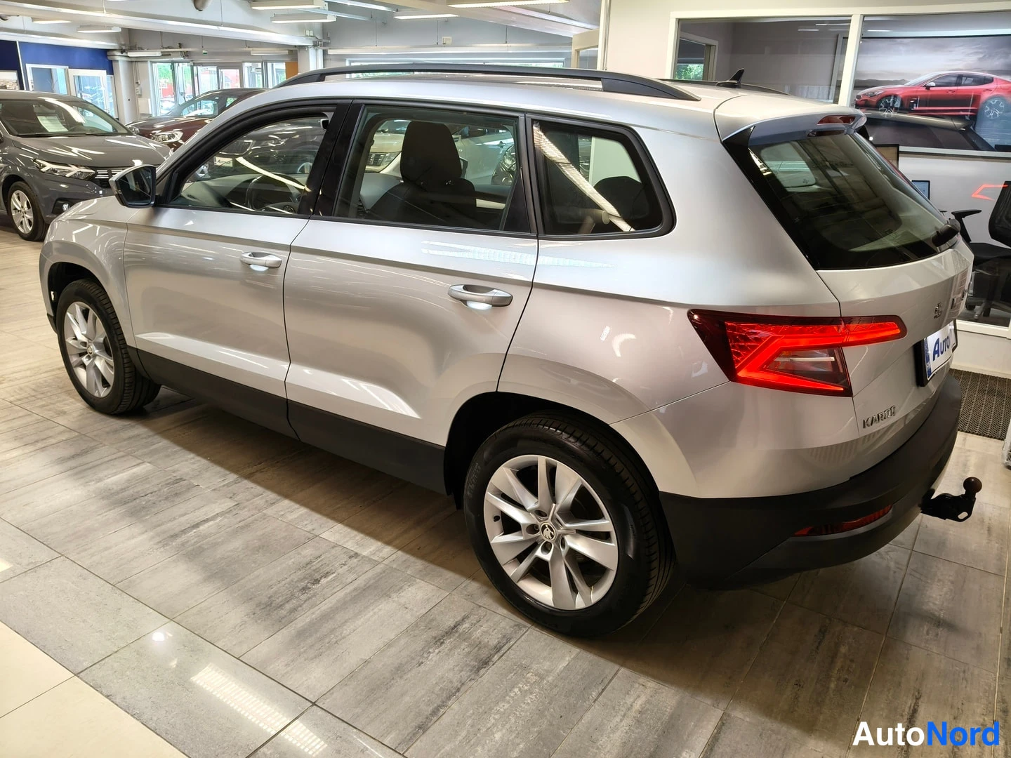 Takaviisto: Hopeanvärinen Skoda Karoq takaosa ja vasen sivu kolmannesnäkymässä autosalissa, perävaunukytkin näkyvissä