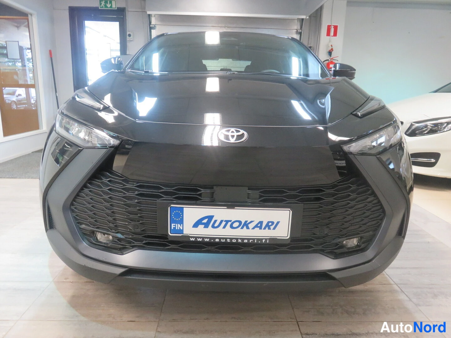 Edestä: Mustan auton etupää myyntitilassa, etupuskurin etualalla valkoinen AUTOKARI-rekisterikilpi