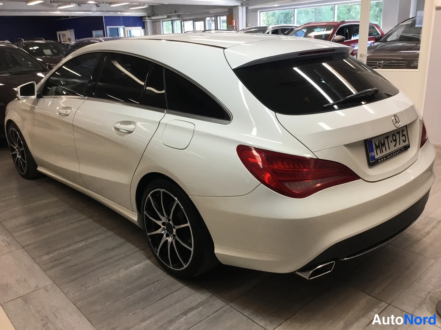 Takaviisto: Rein valkoinen farmarimallinen Mercedes-Benz henkilöauto sisätiloissa, näkyvät takavalo, takapuskuri ja vasemman sivun ovet ja pyörät.