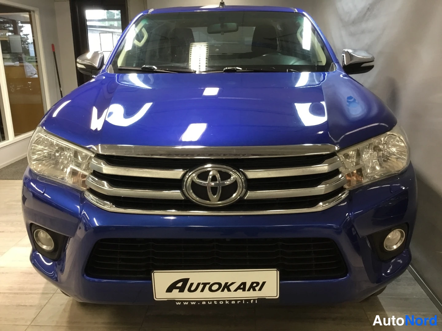Edestä: Sininen Toyota Hilux, etukuva näyttää kromilattiaisen etusäleikön, Toyota-merkin ja sumuvalot.