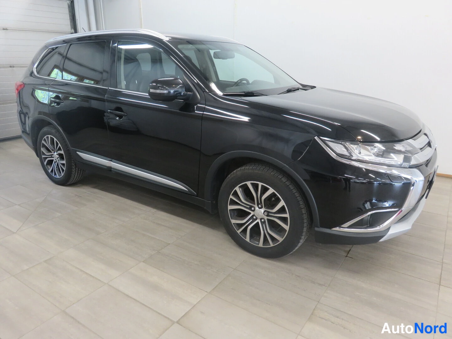 Etuviisto, nokka oikealle: Musta Mitsubishi Outlander -monitoimiauto kuvattu kulmasta etuosaa ja vasenta sivua näkyvin, tilavassa sisätilassa.