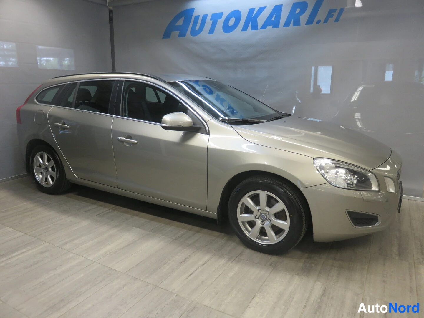 Etuviisto, nokka oikealle: Volvo V60 -farmari sivulta etukulmasta, nokka kuvan oikealla puolella, auton kylki ja etupää näkyvät showroomissa.