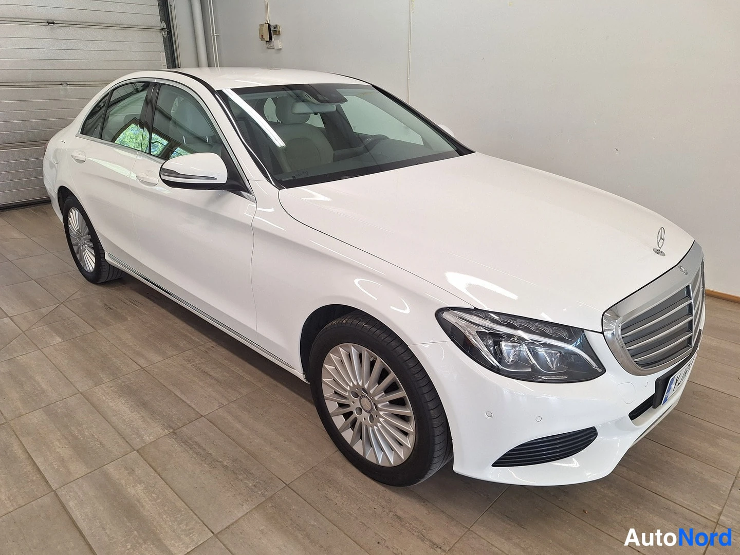 Etuviisto, nokka oikealle: Valkoinen Mercedes-Benz C-sarjan sedan katsottu etuviistosta oikealta, kylki ja keulakoke näkyvät sisätiloissa laatoitetulla lattialla.