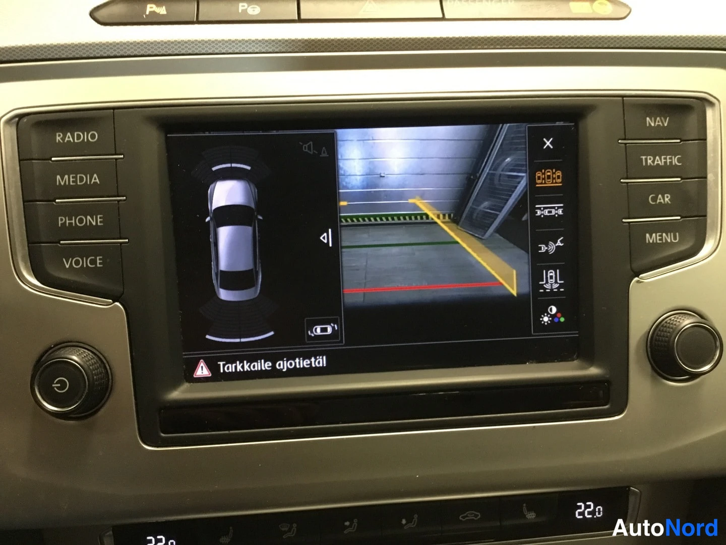 Takaa: Volkswagenin kojelaudan infotainment-näyttö näyttää peruutuskameran kuvan ja pysäköintiruudukon. Ruudulla varoitus 'Tarkkaile ajotietä!'.