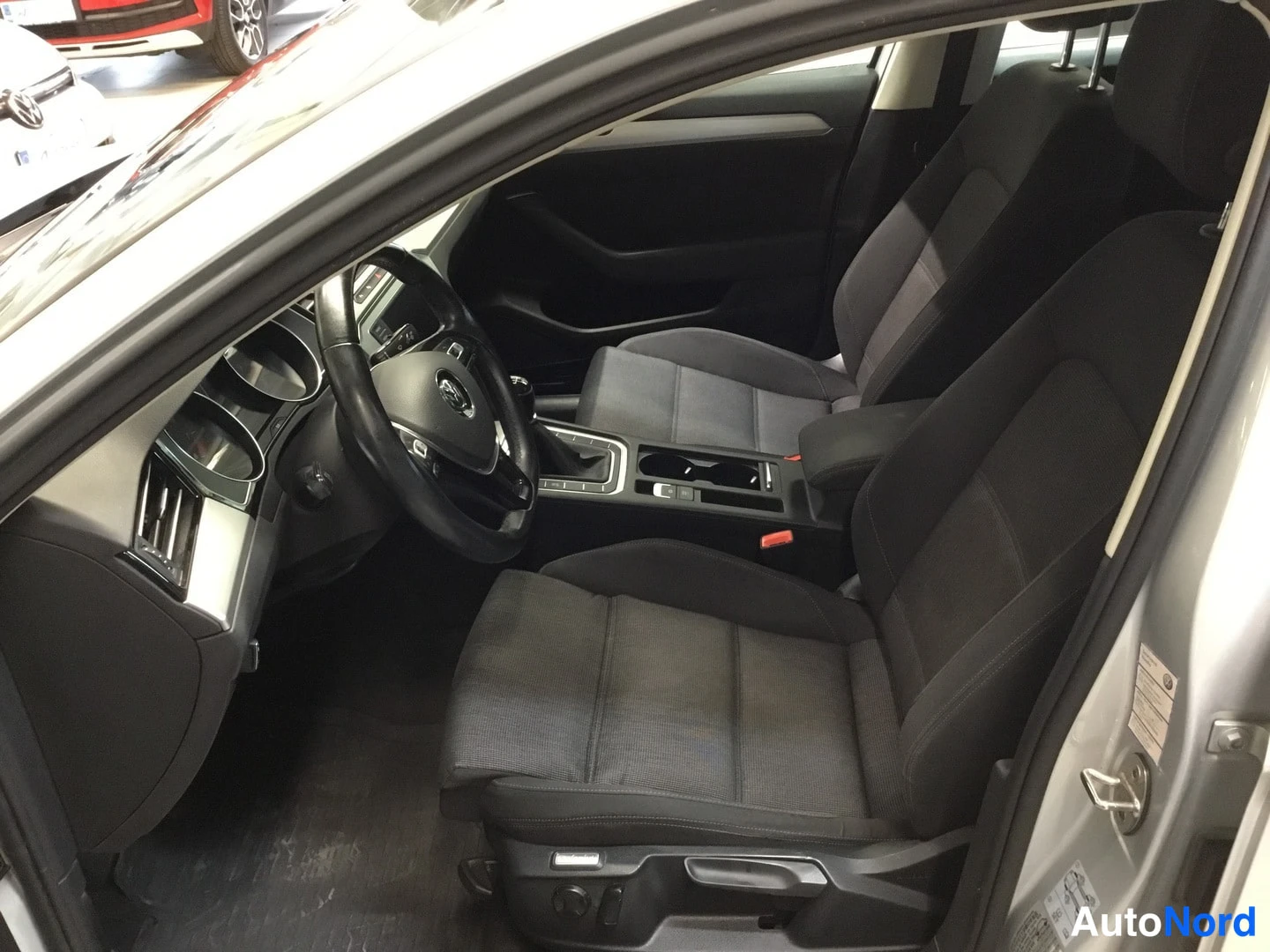 Muu: Näkymä auton sisälle avatusta ovesta: tummat kangasistuimet eturivissä, ohjauspyörä ja keskikonsoli; korin hopeanharmaa pinta näkyy ovenkarmista.