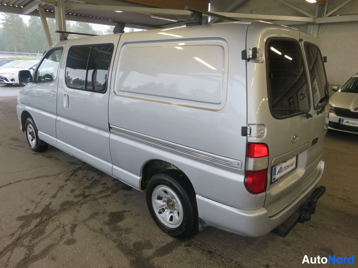 Takaviisto: Hopeanharmaa Toyota Hiace -pakettiauto takaviistosta oikealta kuvattuna; pitkä runko, sivuikkunat ja kattoteline näkyvät.