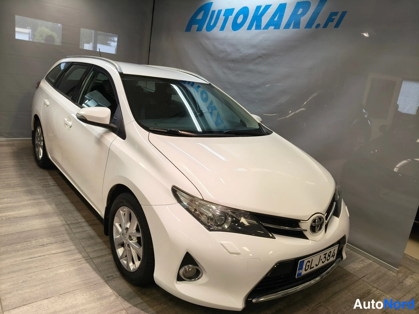 Etuviisto, nokka oikealle: Etukulmasta valokuvattu valkoinen Toyota Auris Touring Sports -farmariauto näytteenottohallissa, rekisteritunnus.