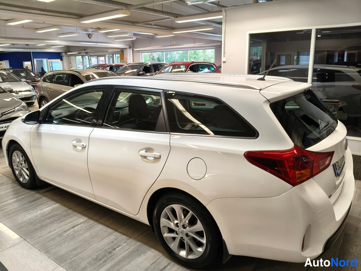 Takaviisto: Takakulmasta valokuvattu valkoinen Toyota Auris Touring Sports -farmariauto autosalon lattialla, punaiset takavalot näkyvissä.