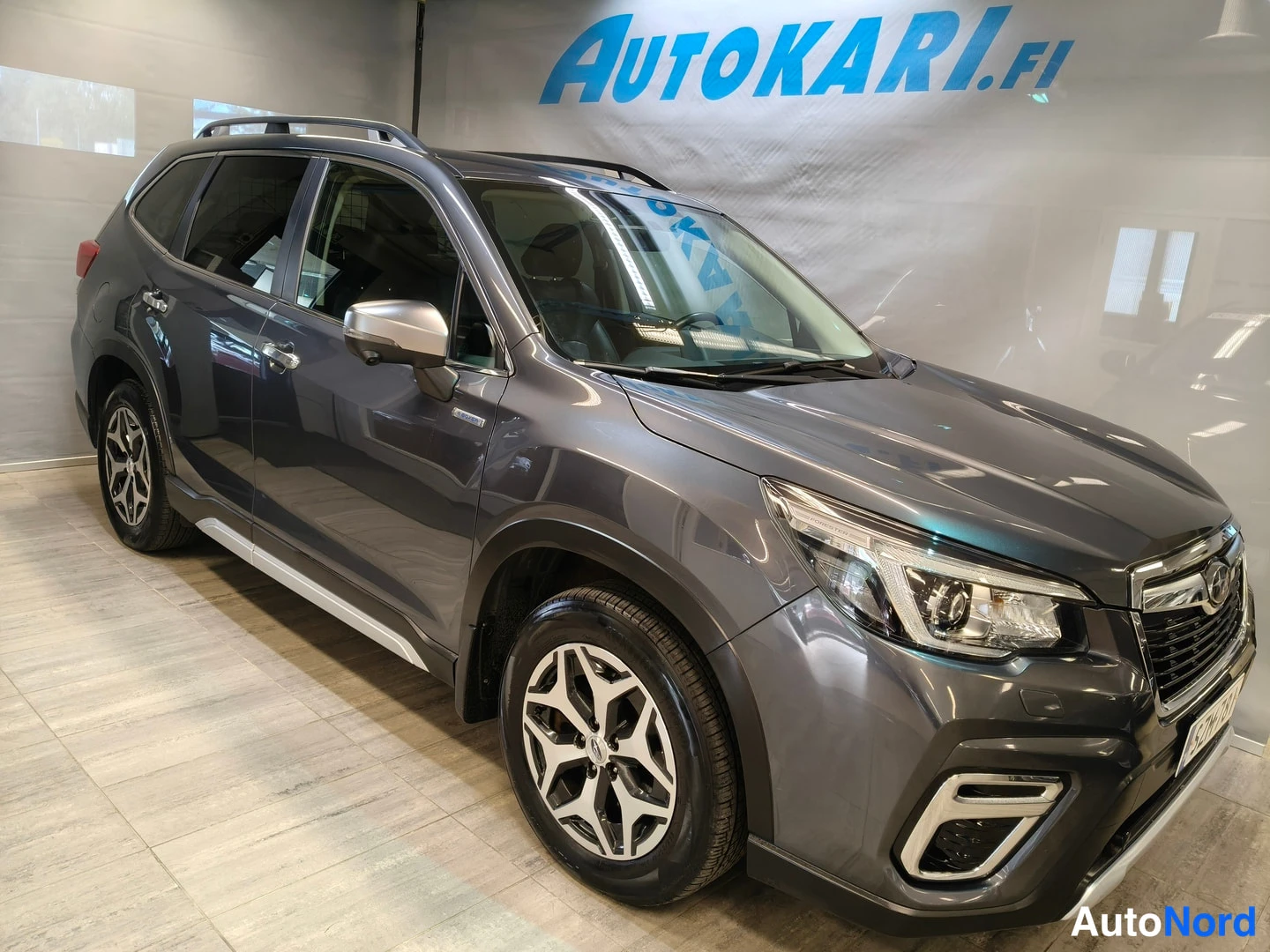 Etuviisto, nokka oikealle: Tummanharmaa Subaru Forester -maasturikuva frontaalista kolmasosaa oikealta, koko auto näkyy sisätiloissa.
