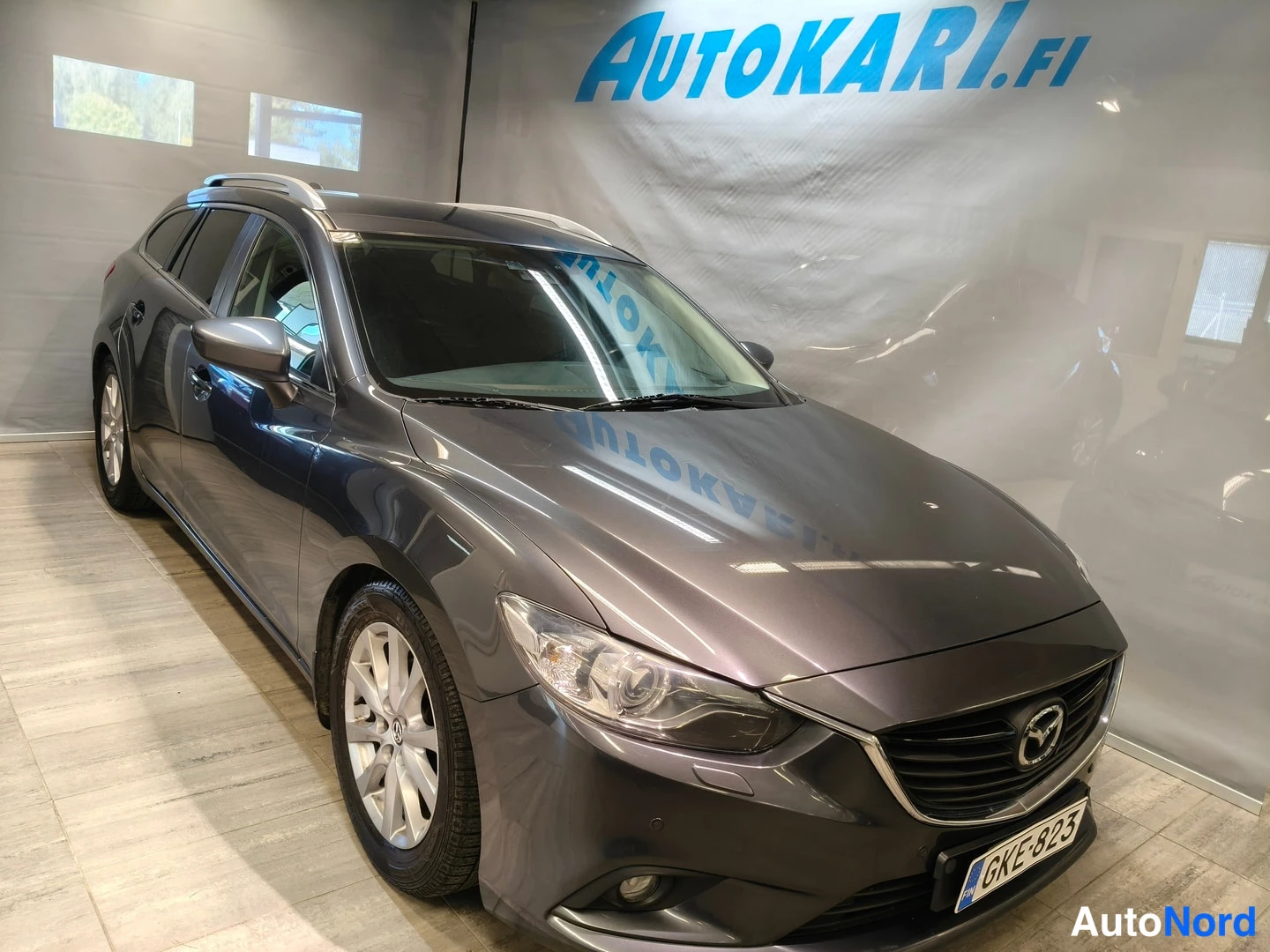 Etuviisto, nokka oikealle: Tummanharmaa Mazda6-farmari etuviistossa oikealta kuvattuna, etupanssari ja kylki näkyvät, rekisteritunnus.