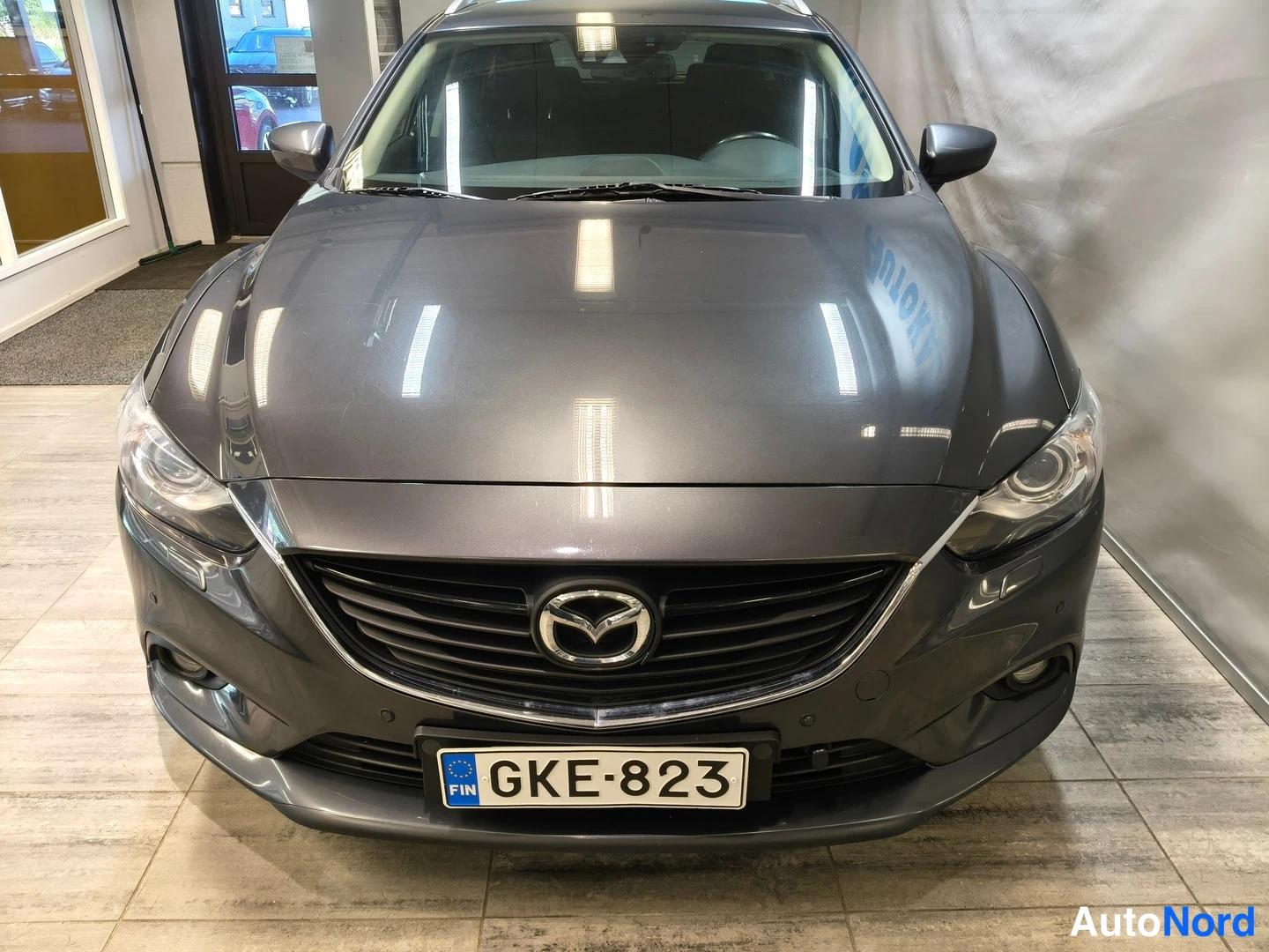 Edestä: Tummanharmaan Mazda6-farisin etupää suoraan edestä, etugrilli ja Mazda-tunnuksella, rekisteritunnus.