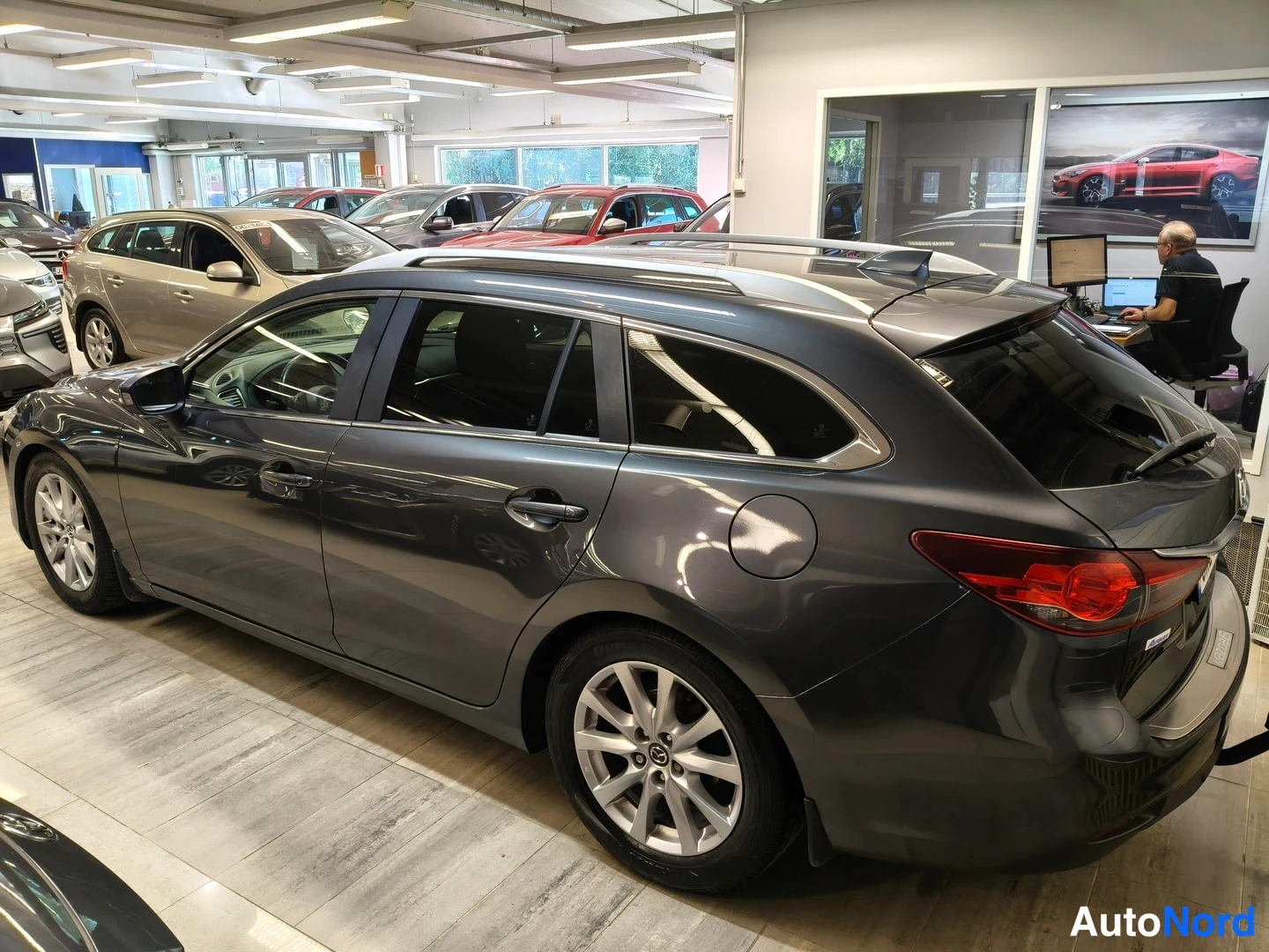 Takaviisto: Tummanharmaa Mazda6-farmari takaviistosta vasemmalta, kylki ja takaosa näkyvät, kattotanko ja peräruuven vuototassu.