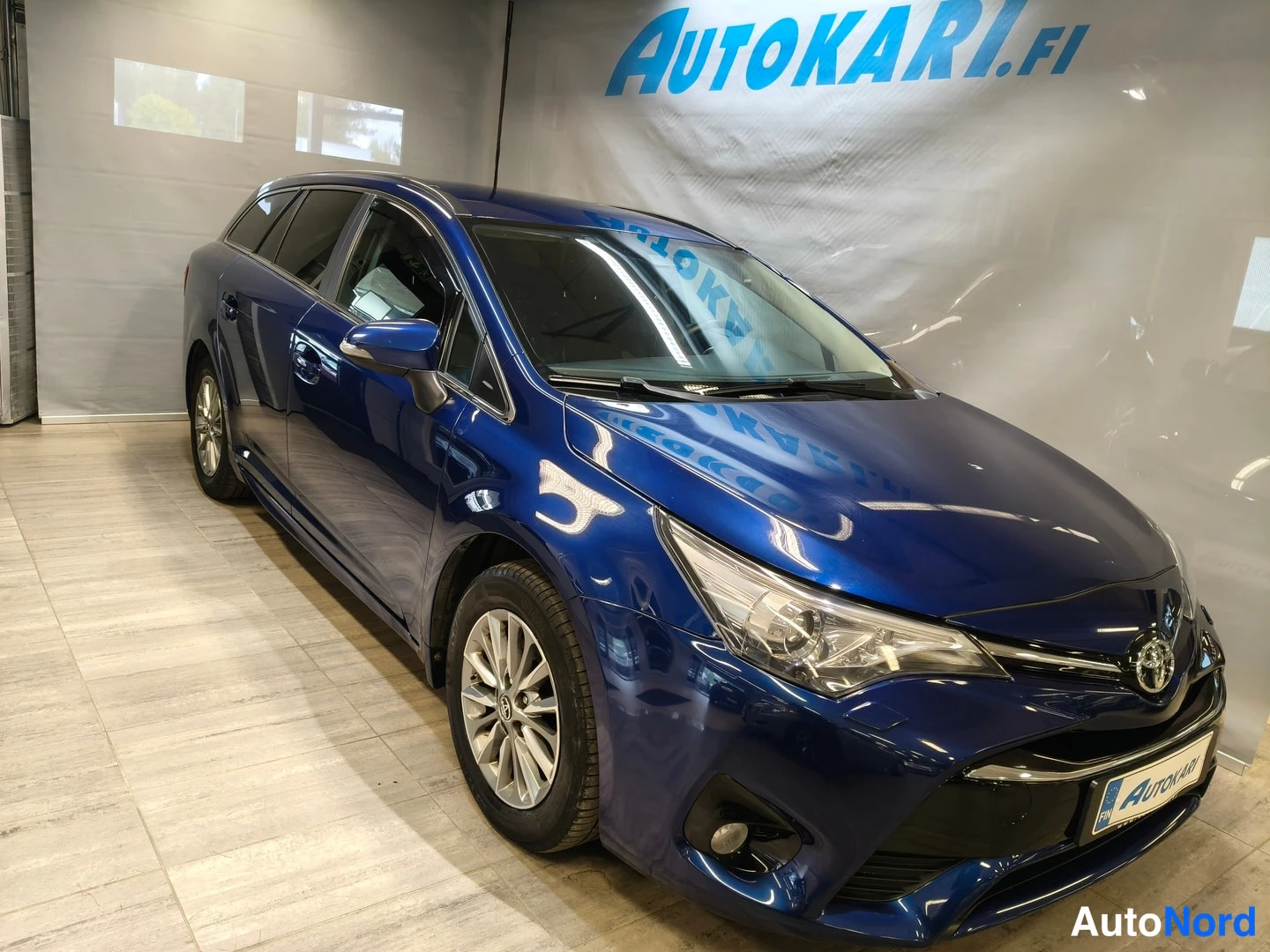Etuviisto, nokka oikealle: Sininen Toyota Avensis -farmariauto etukulmasta oikealta nähtynä,Autokari.fi-tunnuksen alla sisätiloissa.
