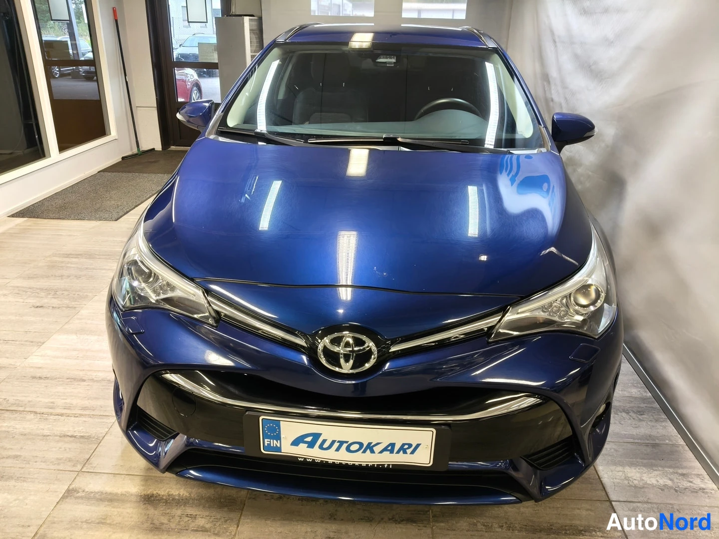 Edestä: Toyota Avensisin etunäkymä: koivärikilpi AUTOKARI-tekstillä ja Toyota-merkki konepellin alla.