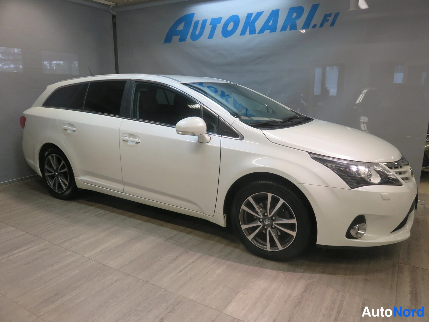 Etuviisto, nokka oikealle: Toyota Avensis -farmari valkoisena etuvasemmalta kuvattuna sisätiloissa.