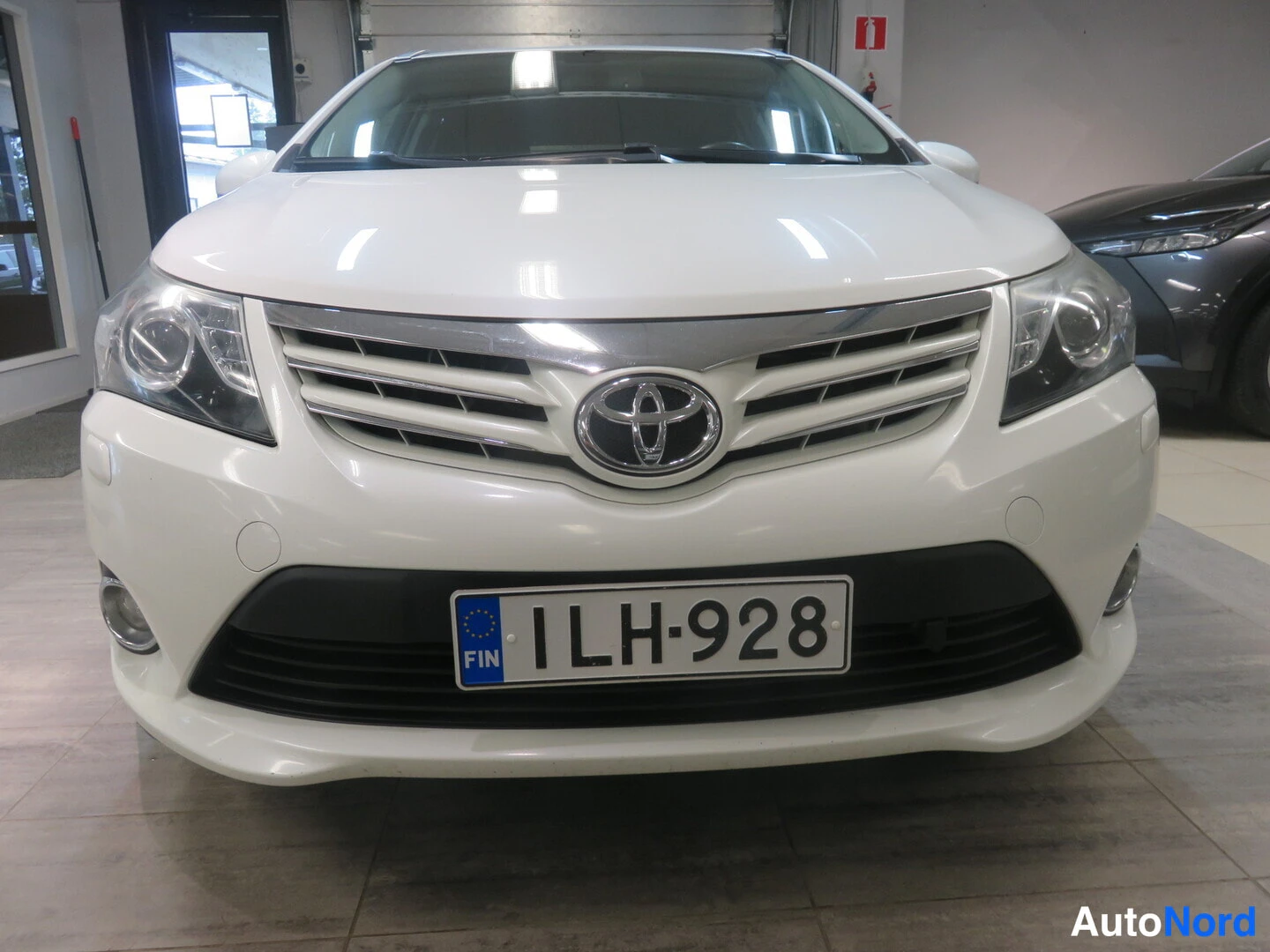 Edestä: Valkoisen Toyota Avensisin keulakuva suoraan edestä, rekisteritunnuksella.